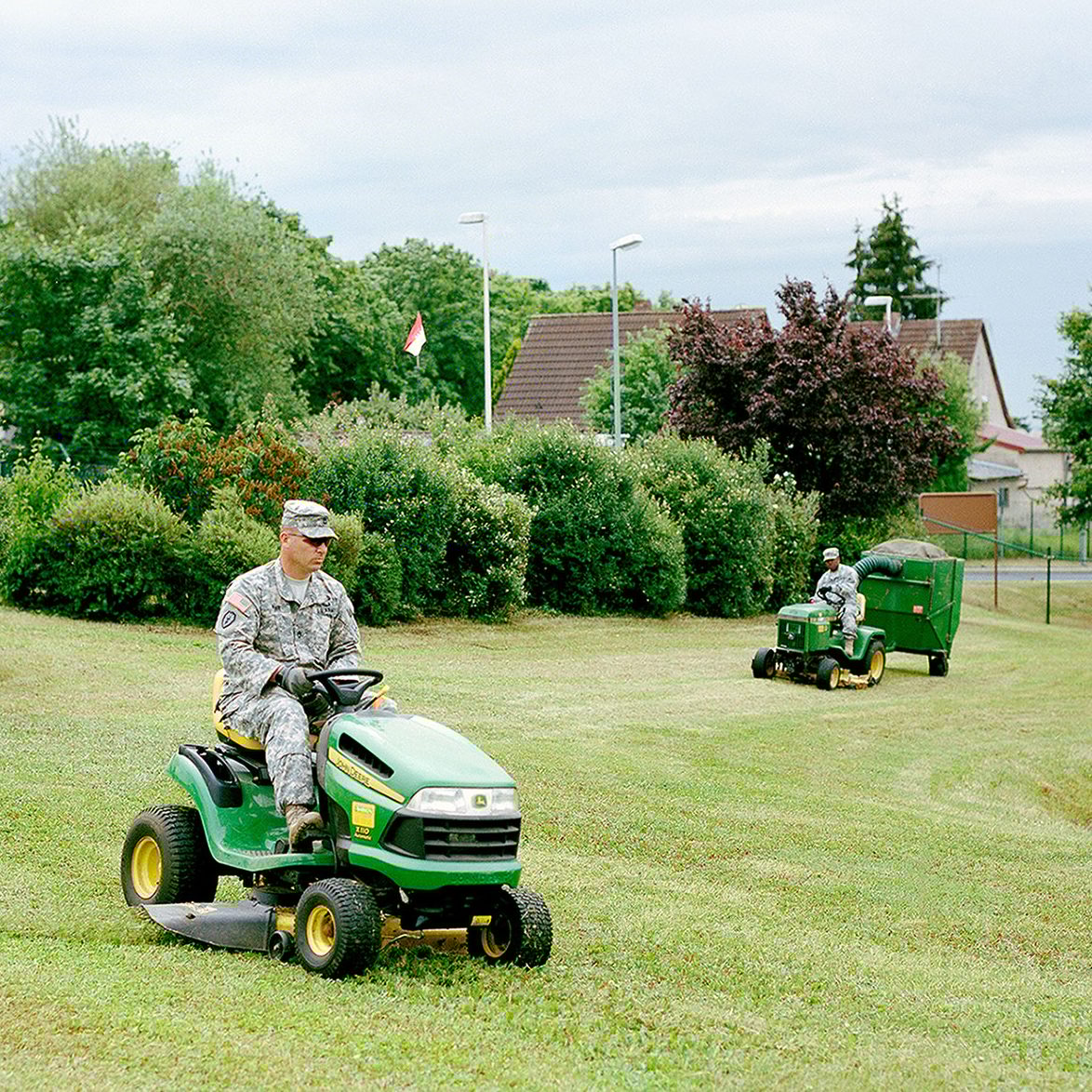 Military personnel mow a lawn with two John Deere riding mowers.