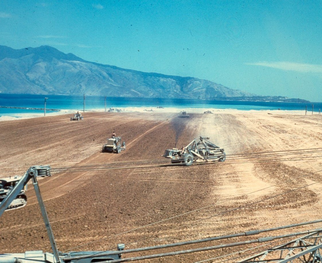 Heavy equipment working a large field by the ocean with mountains; power lines in foreground.
