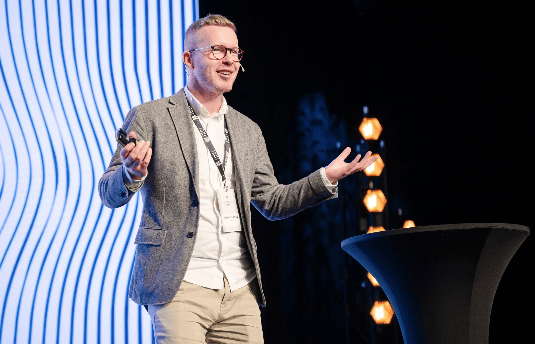 A man in glasses and a grey jacket speaks on stage with a blue patterned backdrop and illuminated stand.