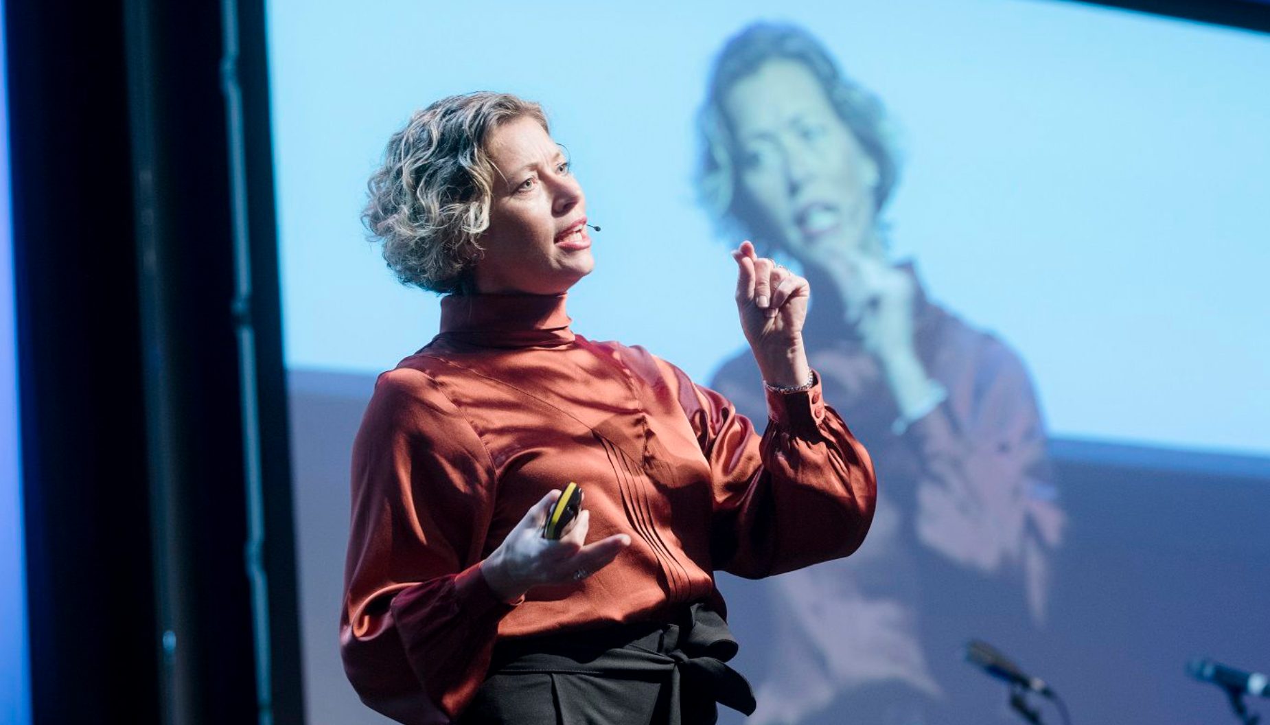 A woman with short curly hair in a red blouse presents on a stage with a blurred screen behind her.