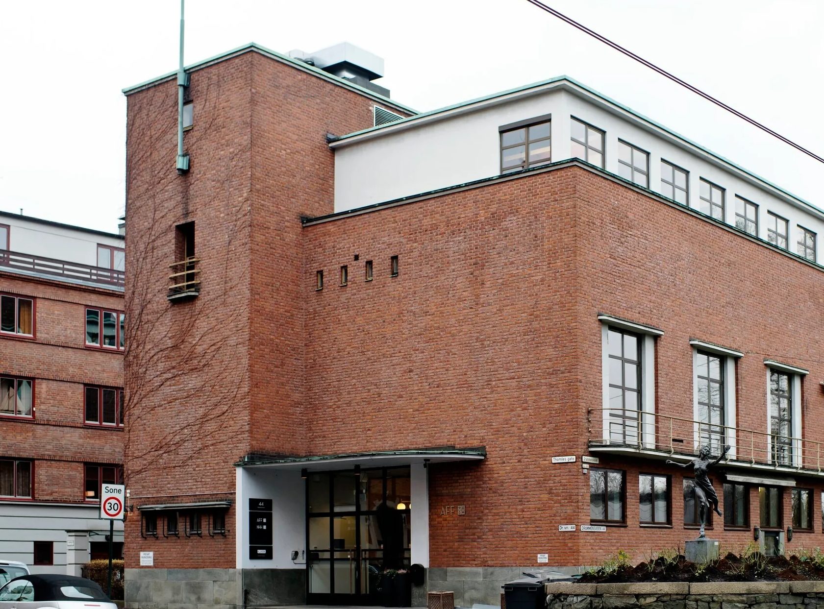 A red brick building with white accents, large windows, and a modern design. Ivy climbs one wall, a statue stands outside.
