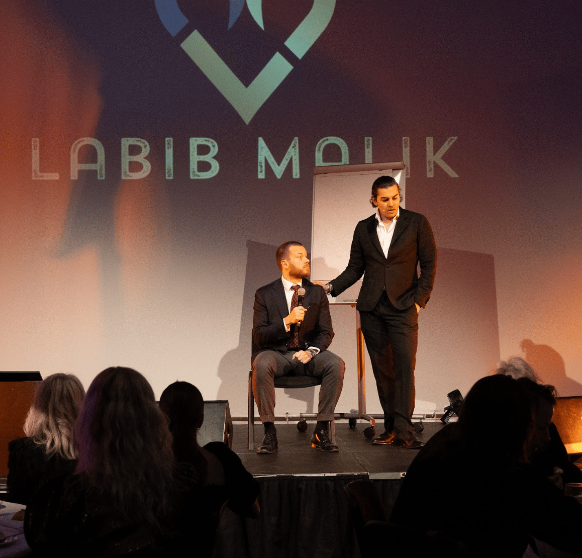 Two speakers on stage: one seated with a mic, one standing. "LABIB MALIK" projected. Audience in view.