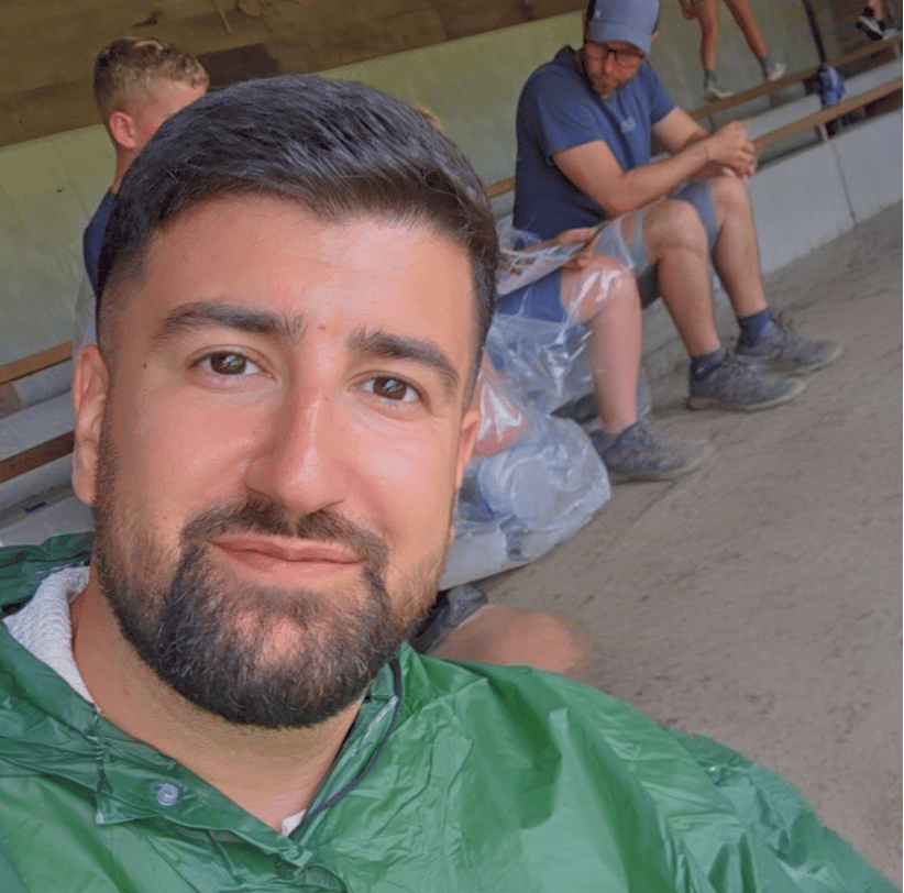 Close-up selfie of a smiling man in a green raincoat, with people sitting in the blurred background.