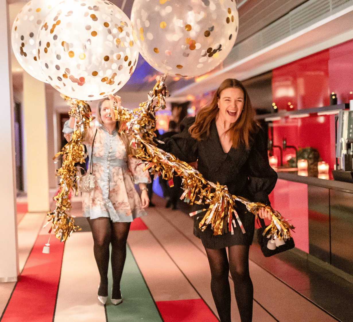 Two women celebrating with confetti balloons and gold streamers, laughing in a vibrant setting.