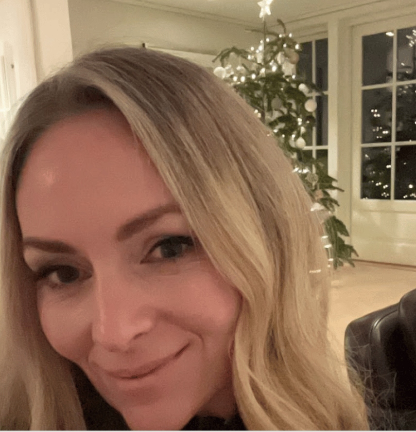 Close-up of a smiling blonde woman with a decorated Christmas tree behind her.