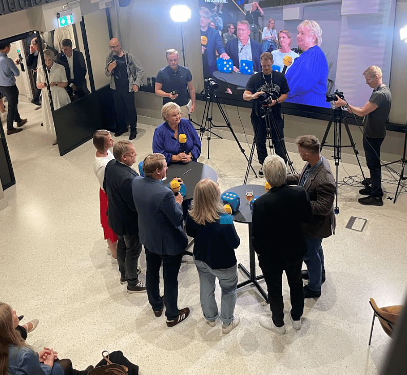 A woman speaks into microphones, surrounded by a film crew, audience, and a large screen showing the event.