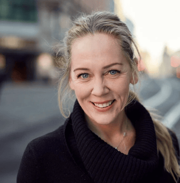 Smiling blonde woman with blue eyes in black top, outdoor setting.