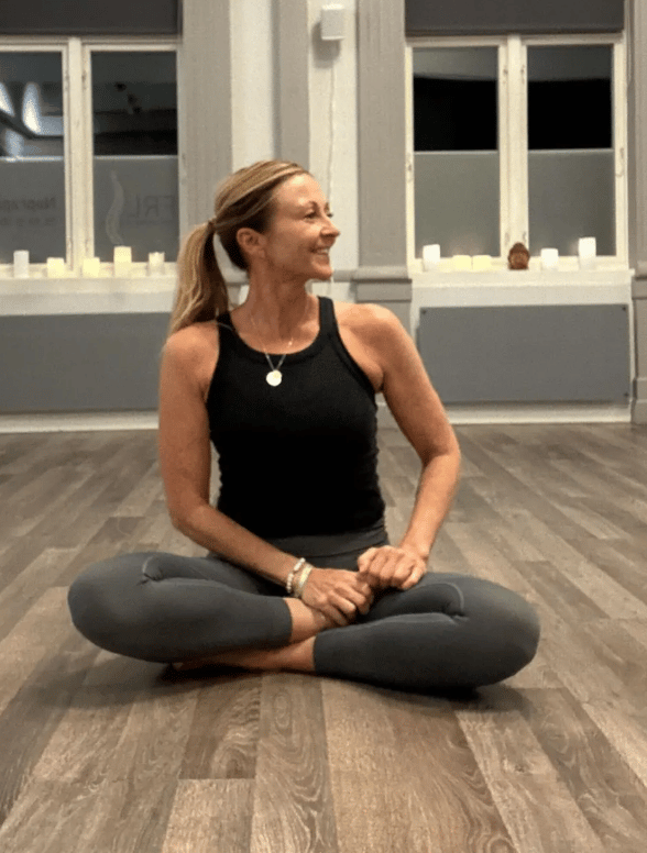 Smiling woman in black top and grey leggings, sitting cross-legged on a wooden floor.