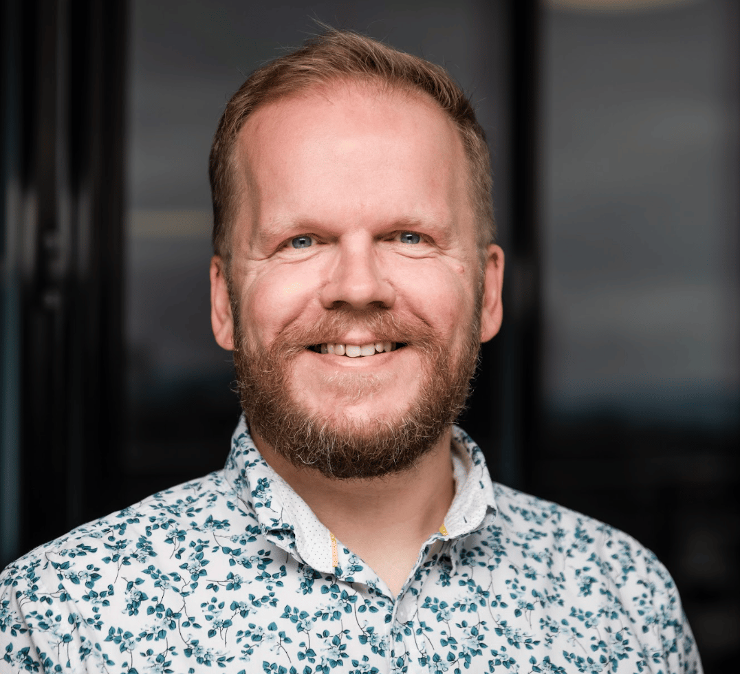 Smiling bearded man with blue eyes in a patterned shirt.