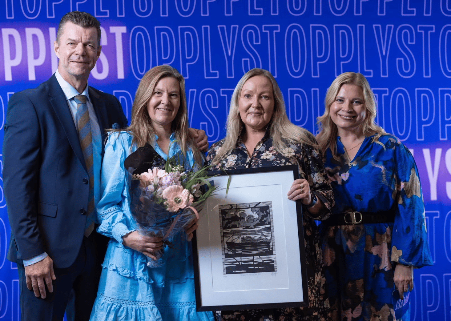 Man and three women pose with flowers and framed art against a blue background.