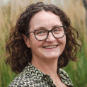Smiling woman with curly brown hair, glasses, and a patterned shirt against a blurred background.