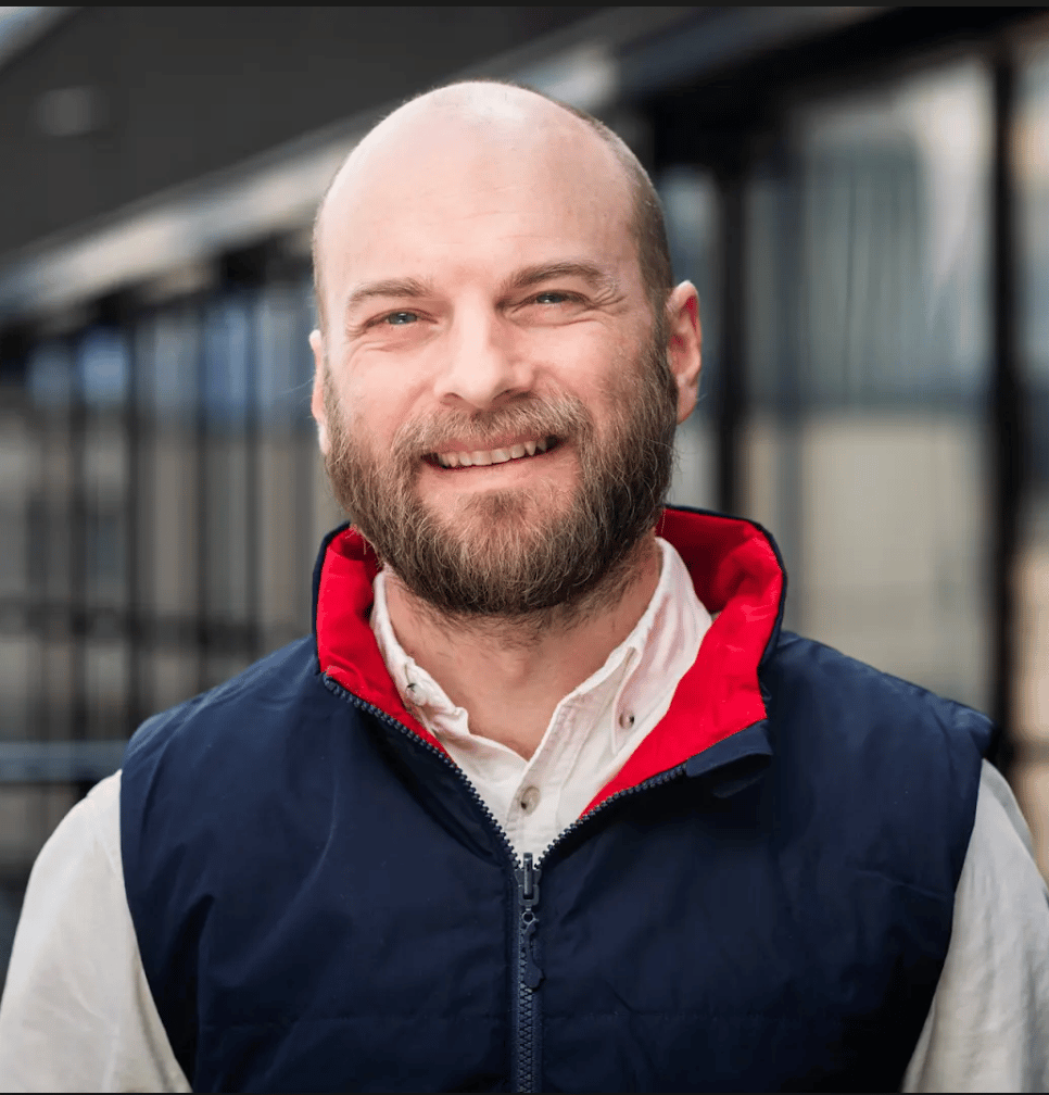 Smiling bald man with beard in navy and red vest.