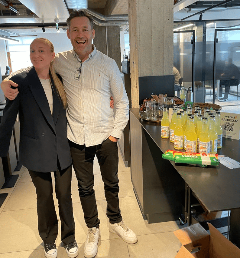 Two smiling people by a table with orange drinks, chocolate, and an "Amedias PÅSKEQUIZ" sign.