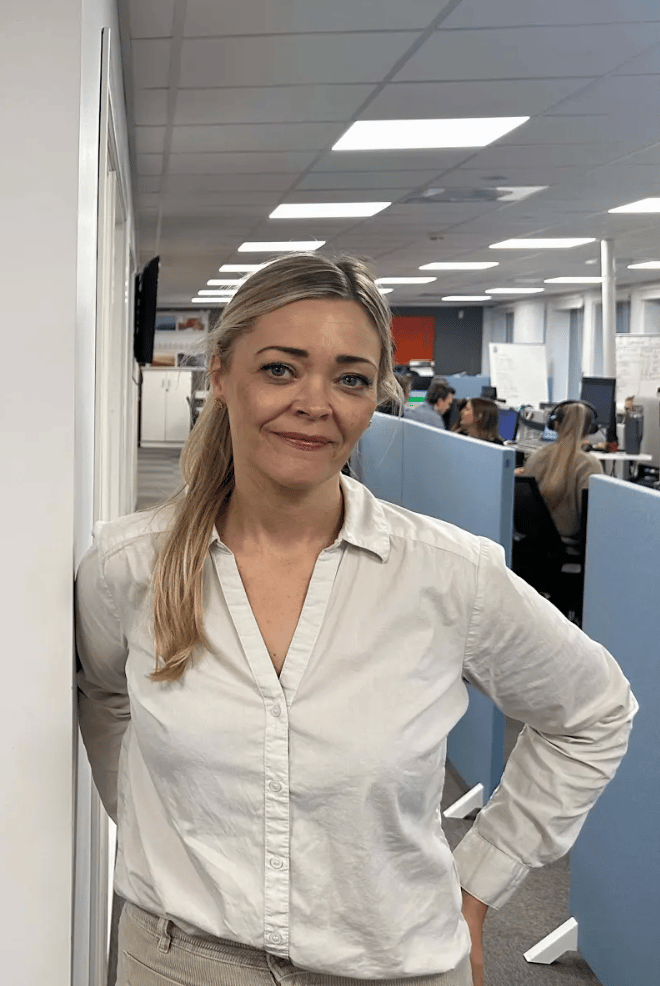 Blonde woman in white shirt smiles in an office, leaning against a wall.