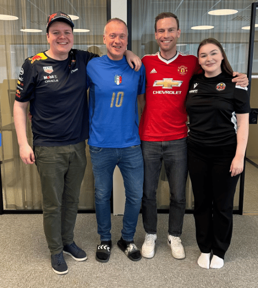 Three men and one woman smiling in sports jerseys: Red Bull, Italy, Manchester United, and a black jersey.