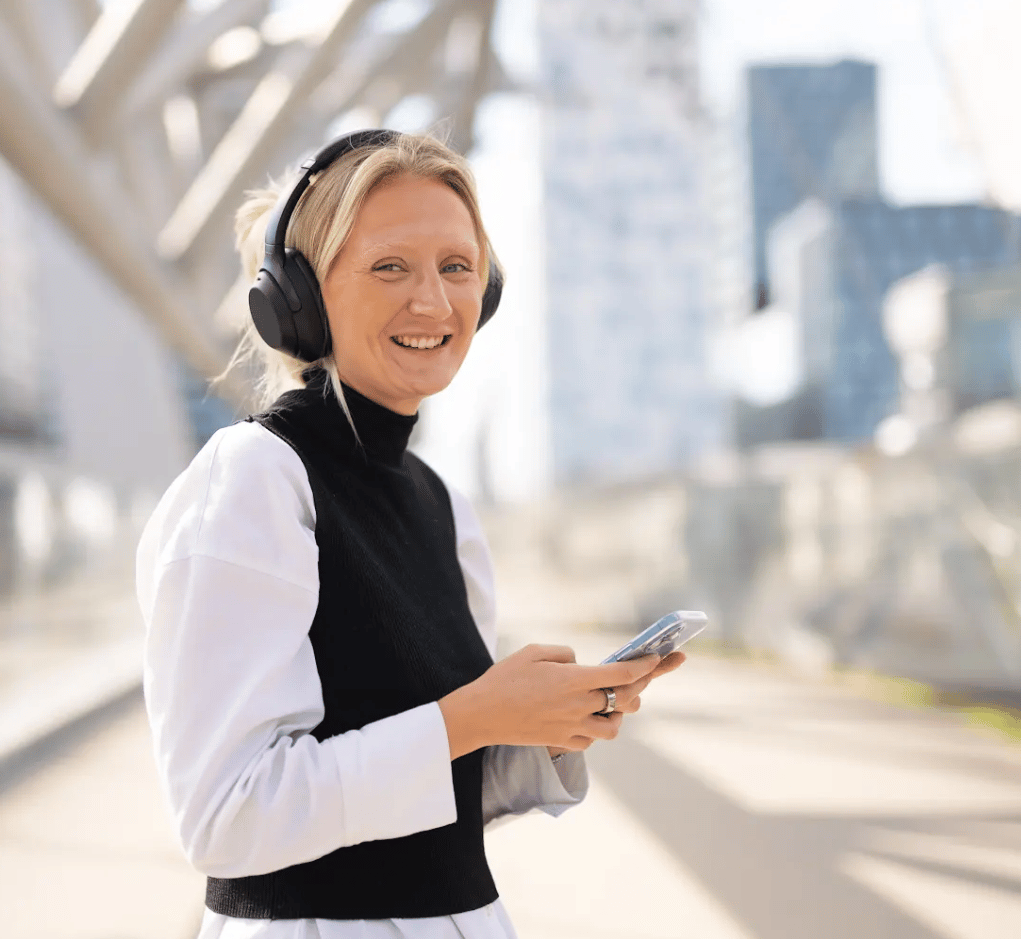 Smiling woman with headphones holding a phone outdoors.