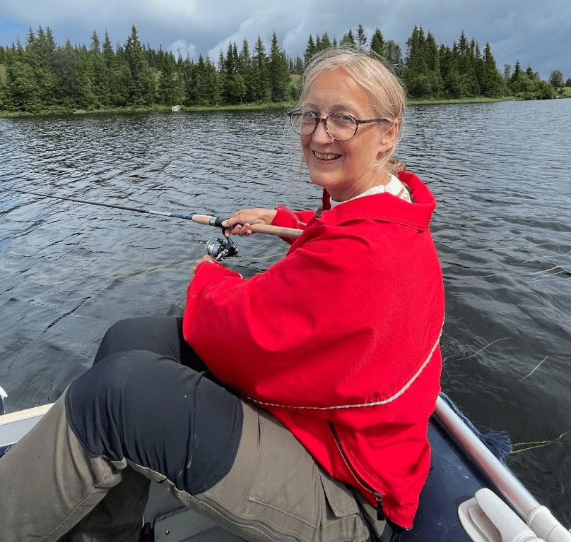 Smiling woman in a red jacket fishing from a boat on a lake with trees.