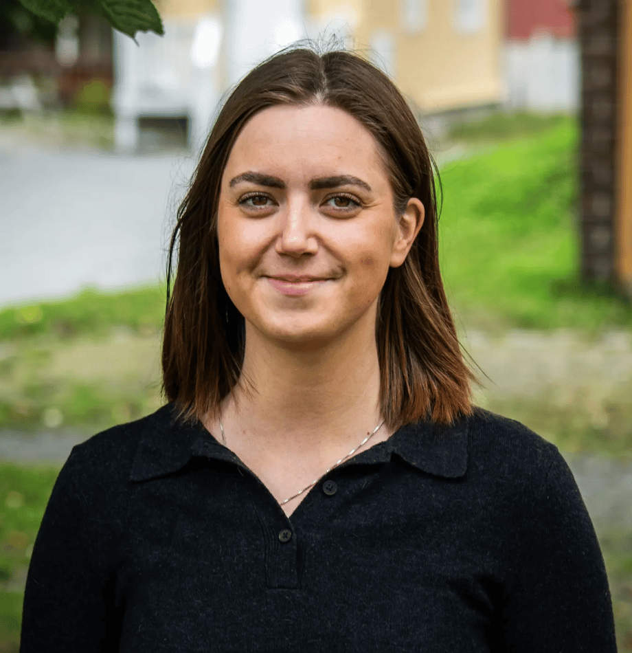 Smiling young woman with brown hair, dark shirt, silver necklace outdoors.