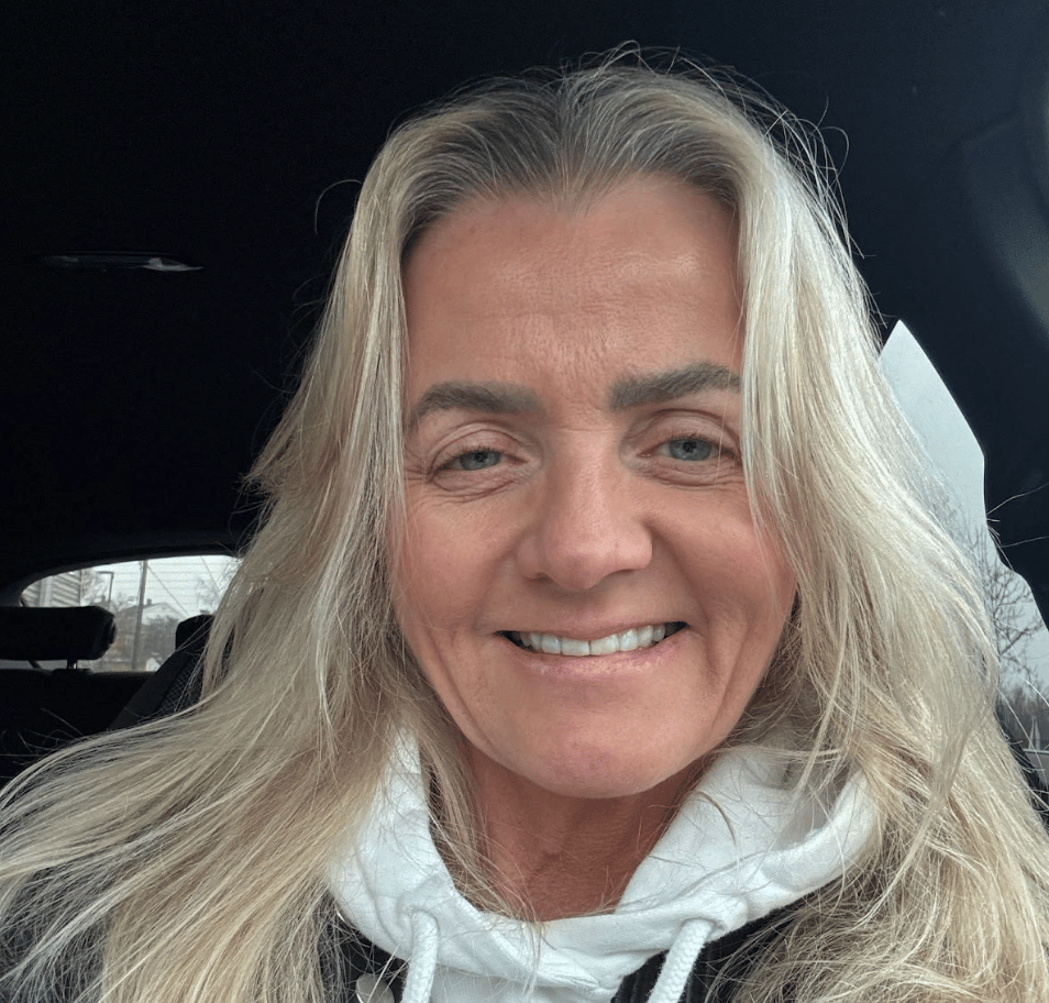 Smiling woman with long blonde hair and a white hoodie, sitting in a car.