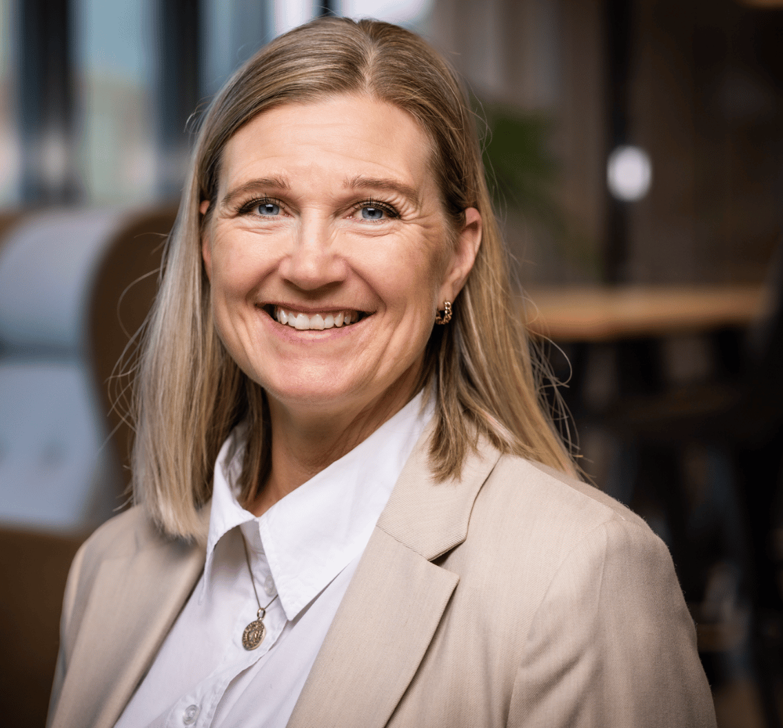 Smiling blonde woman with blue eyes, wearing a beige blazer.