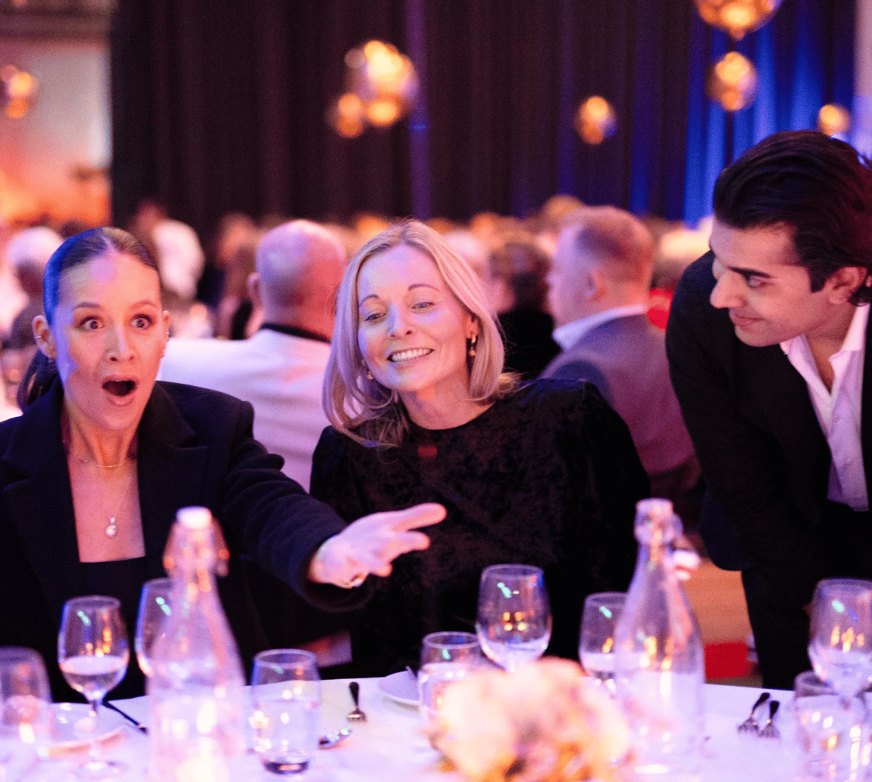 Surprised woman with open mouth and outstretched arm, smiling woman, and man at a formal dinner.