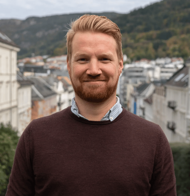 Smiling man with red beard and blond hair. City and hills in background.