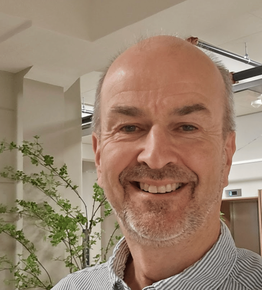 Smiling man with receding hairline and beard, wearing a striped shirt, green plant background.
