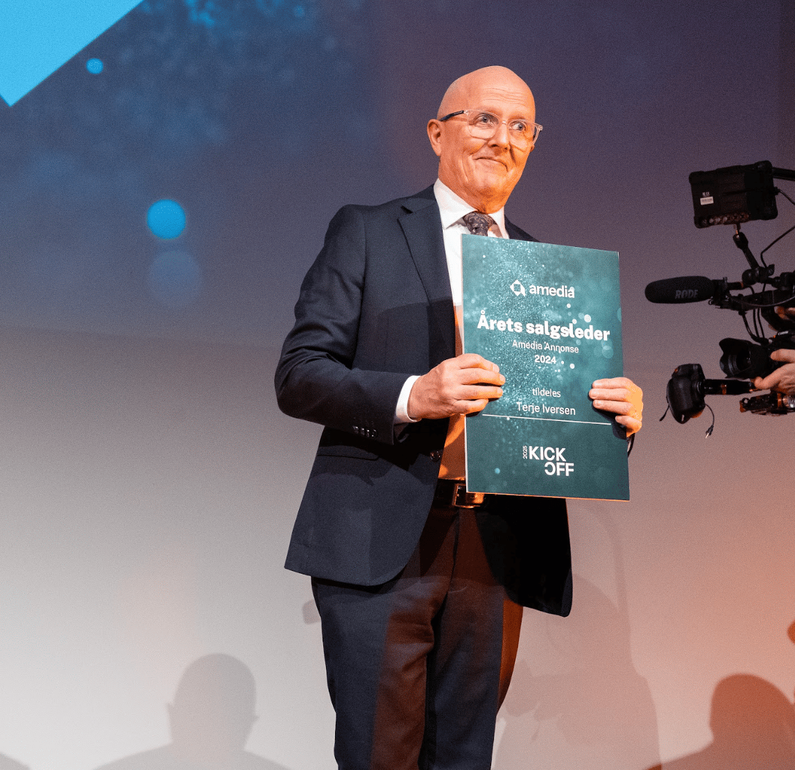 A bald man in a suit and glasses smiles while holding a green "Årets salgsleder" award.