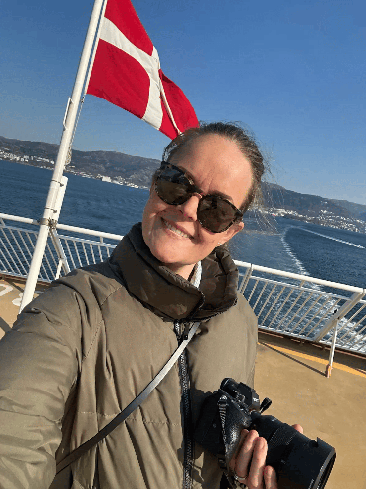 Smiling woman takes a selfie on a ferry with a Danish flag, ocean, mountains, and a camera.