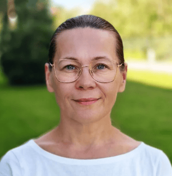 Smiling woman with glasses against a green outdoor background.