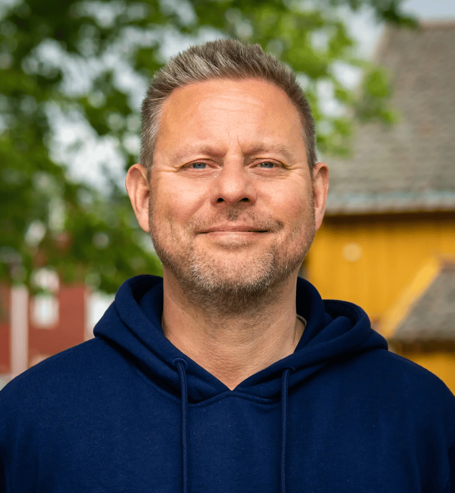 Smiling man with graying hair and beard in a blue hoodie, against a blurred outdoor background.