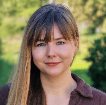 Smiling woman with brown hair, bangs, and blue eyes outdoors.