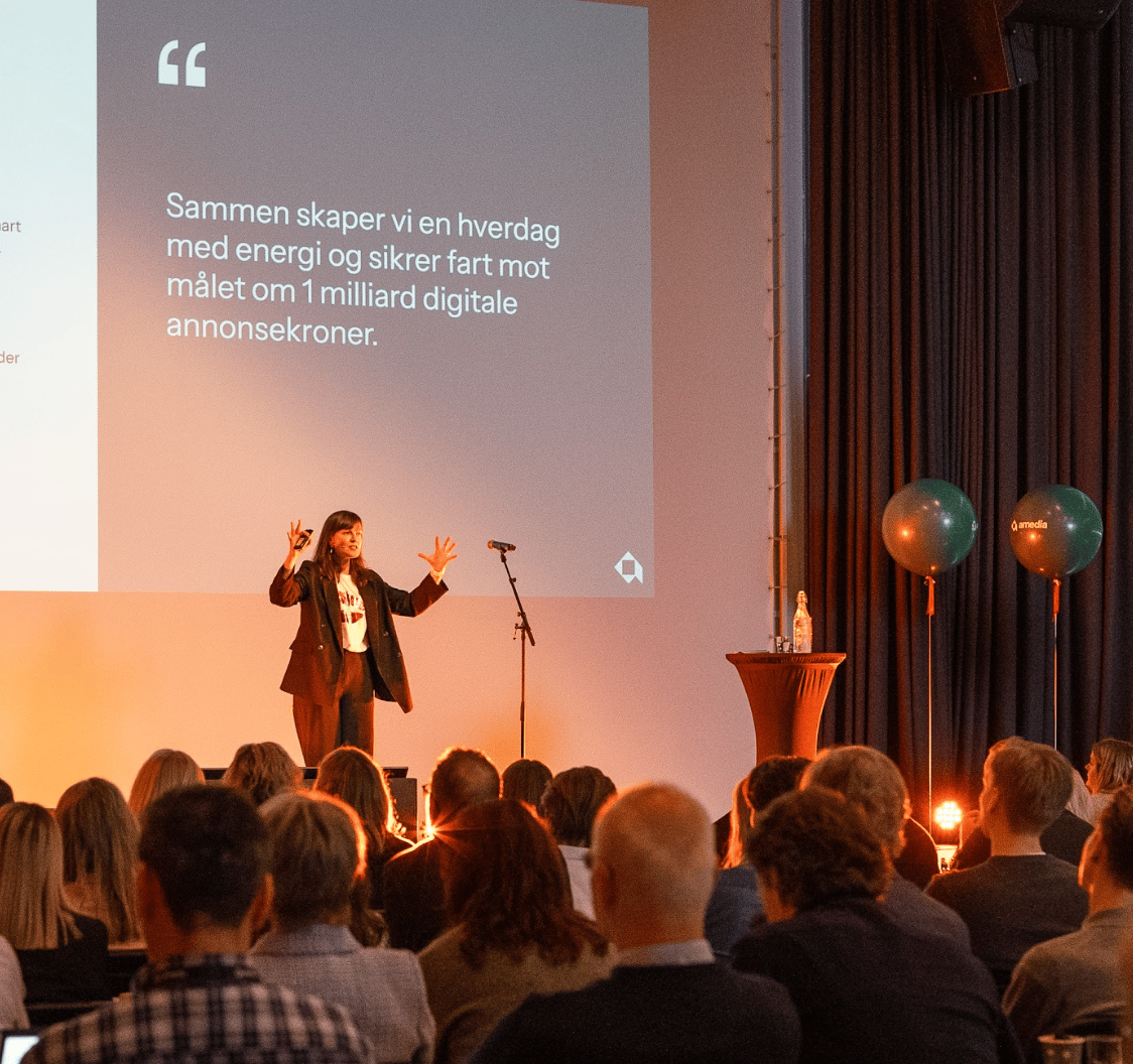 Woman presents on stage to an audience, a large screen with Norwegian text behind her.