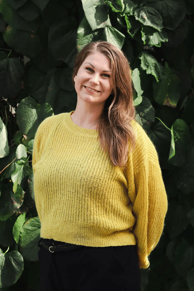 Smiling woman in yellow sweater and black pants against green foliage.
