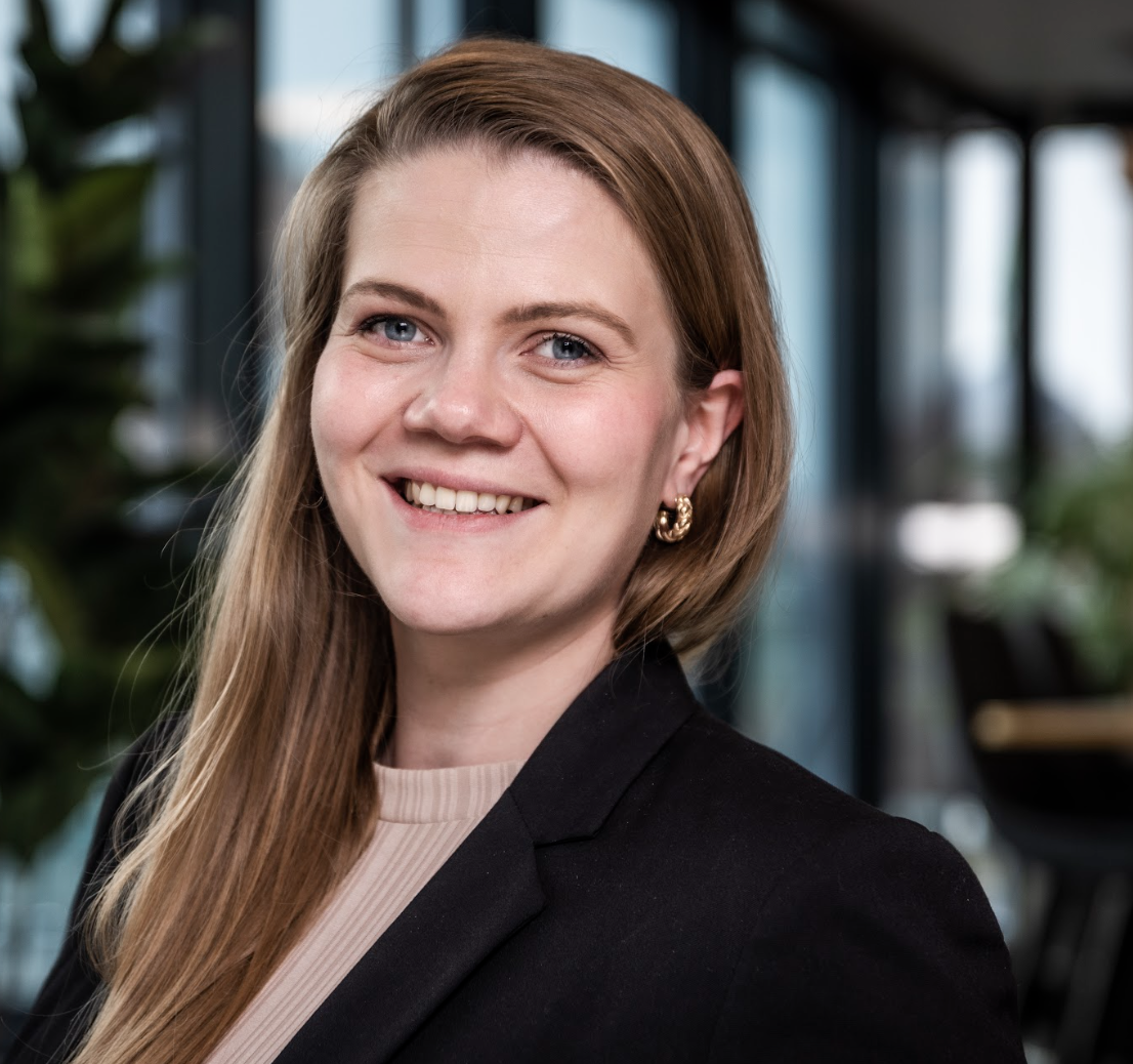 Smiling blonde woman in a black blazer against a blurred office background.