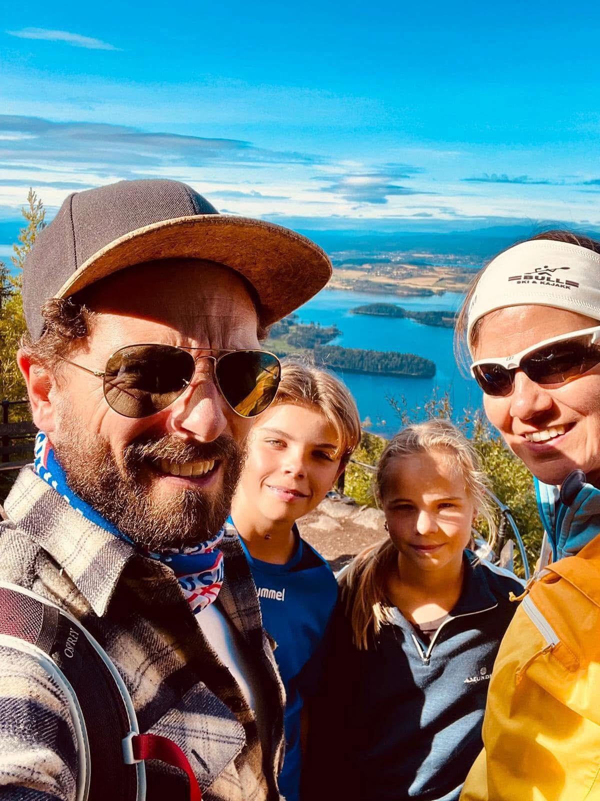 A family of four smiles in a selfie on a mountain peak, overlooking lakes, islands, and a distant landscape.