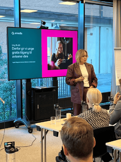 A woman presents in front of a screen about free newspaper access for youth, to an audience.