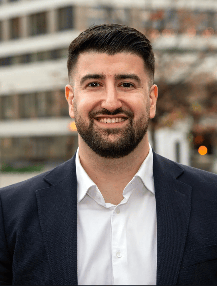 Smiling man with beard, dark suit, white shirt, blurred city background.