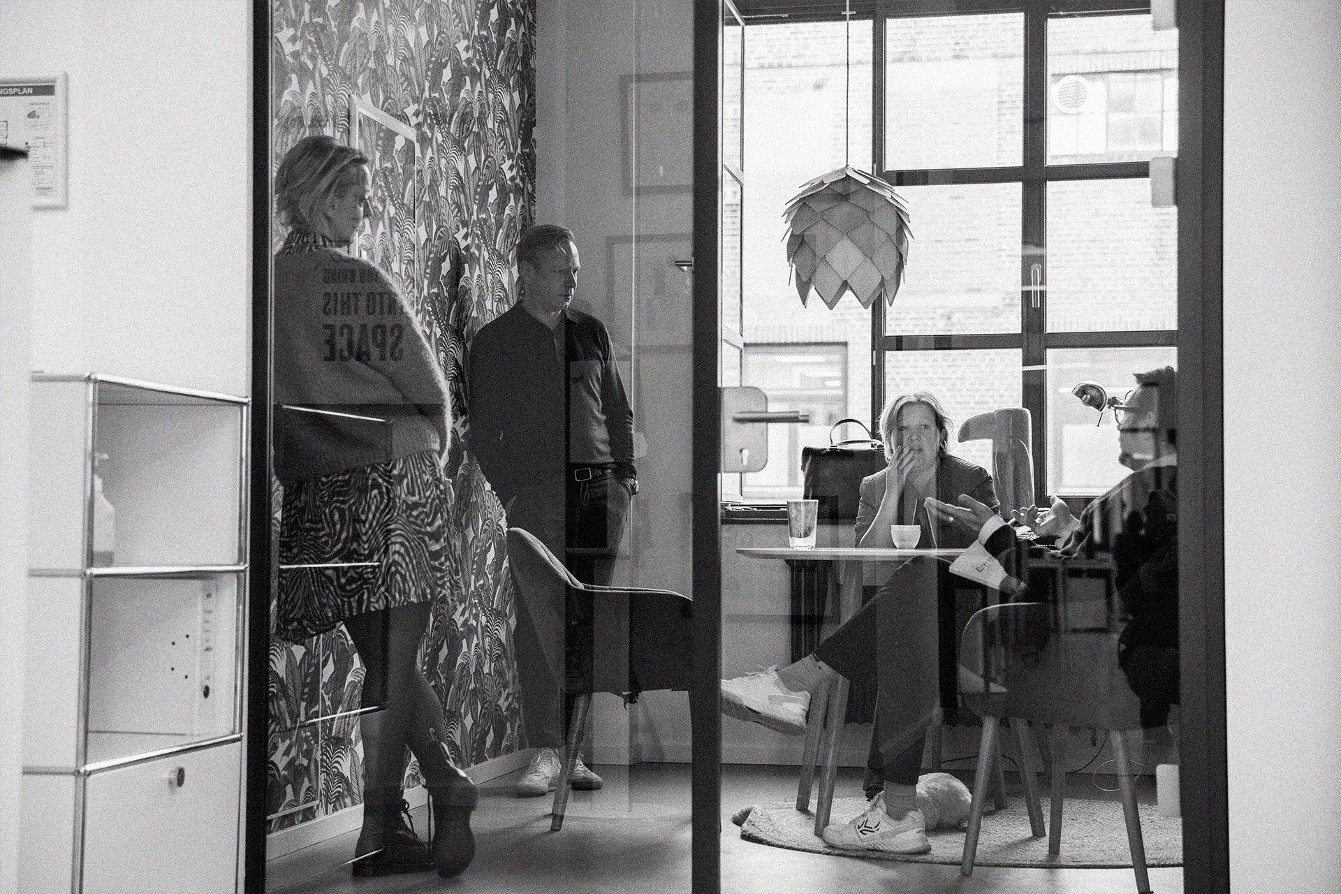 Monochrome office scene with glass partition: people standing are reflected while others are visible through the glass.
