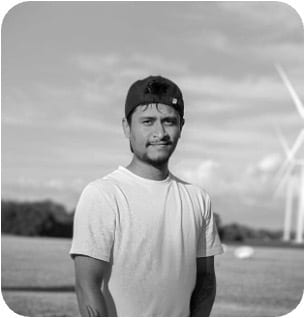 Black and white photo of a young man in a t-shirt and backward cap, standing in a field with wind turbines.