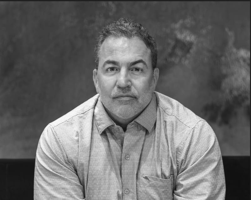 B&W portrait of a man with short hair and beard in a checked shirt, looking at camera.