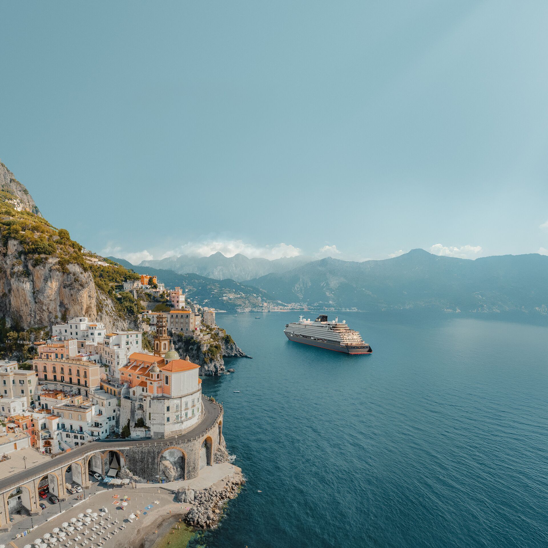 Scenic coastal town built into cliffs, with a cruise ship in the blue sea and distant mountains.