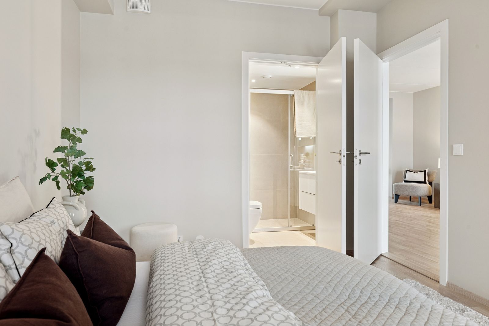 Modern bedroom with bed, plant, and open doors to a bathroom and another room. Neutral tones.