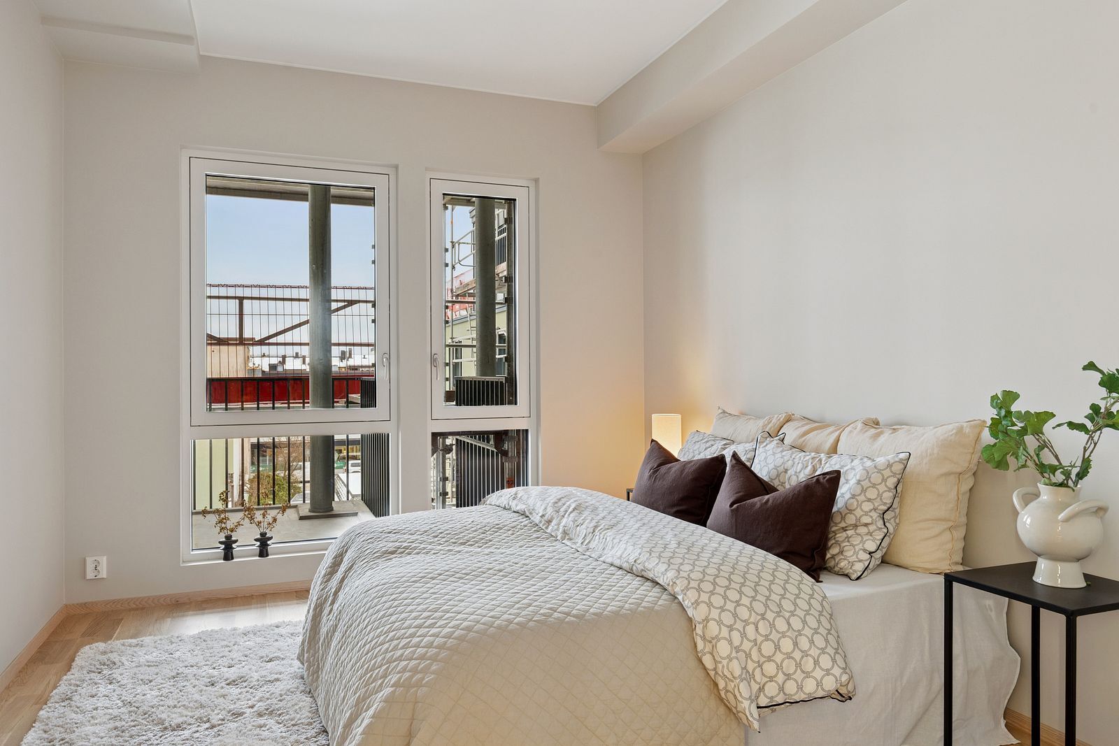 A minimalist bedroom featuring a large bed, fluffy rug, and bright windows with city views.