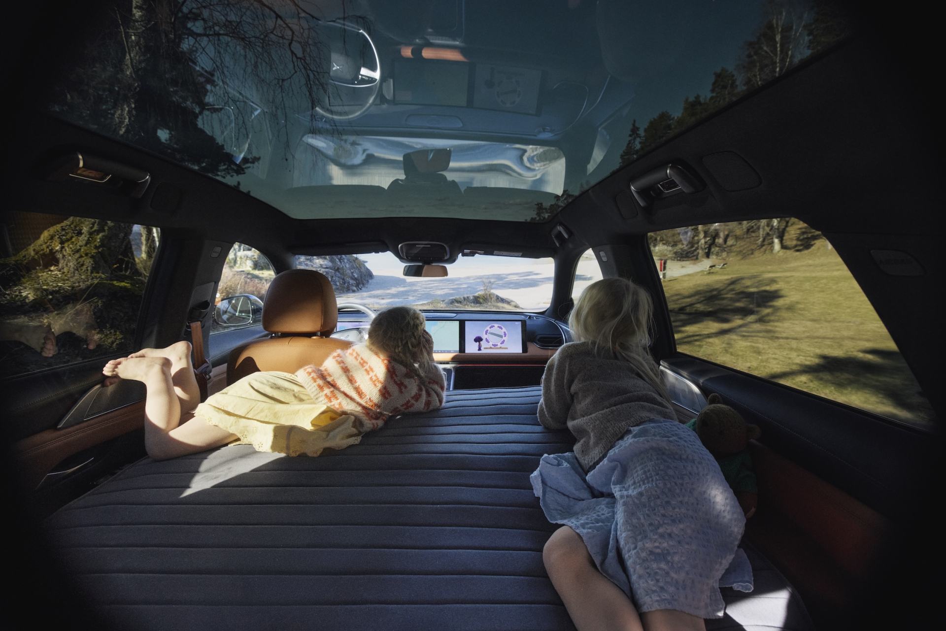 Two children relax on an inflatable mattress in a car with a panoramic roof, looking at screens and outside.
