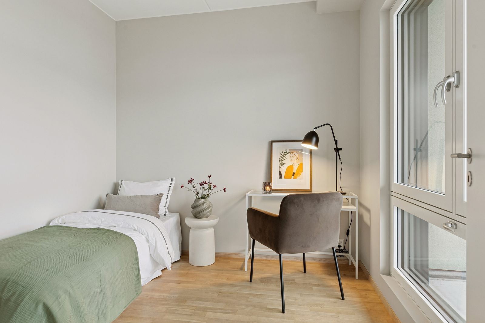 A compact bedroom featuring a single bed, a nightstand, a small desk with a lamp, a chair, and a large window.