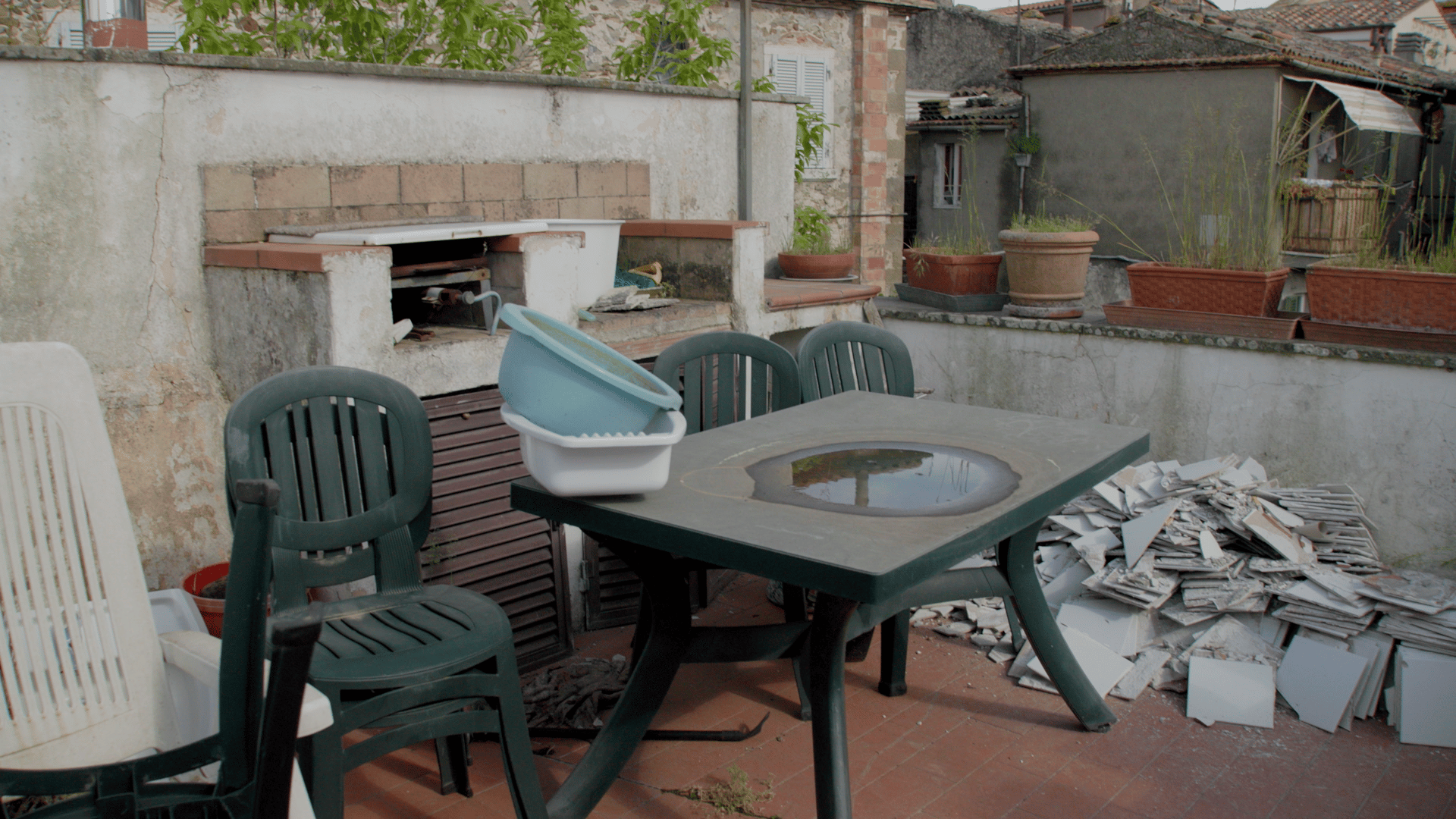 Cluttered rooftop terrace with a green table, chairs, a built-in outdoor kitchen, and broken tiles.