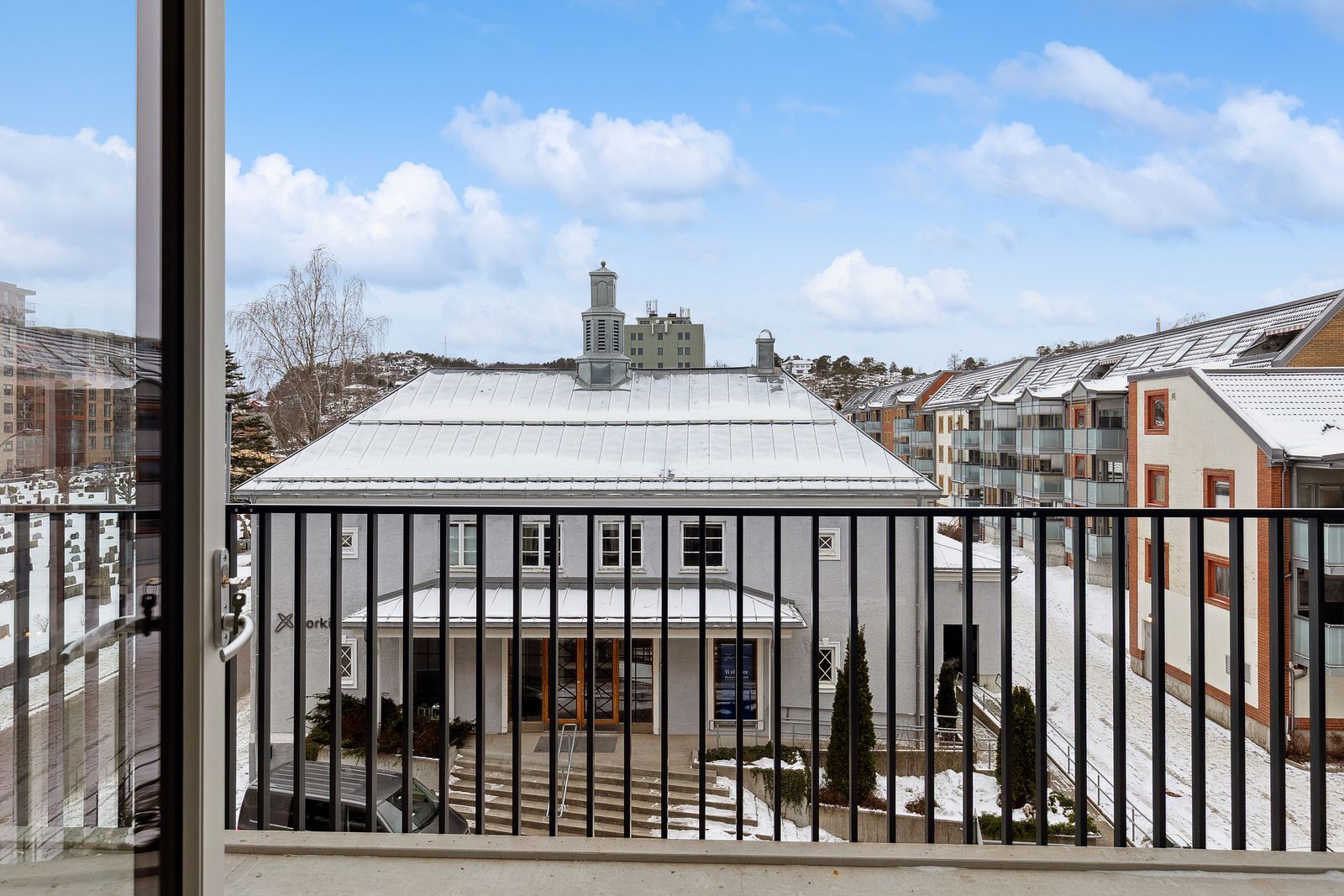 View from a balcony of a snowy urban landscape with a large grey building, distant structures, and a cloudy sky.