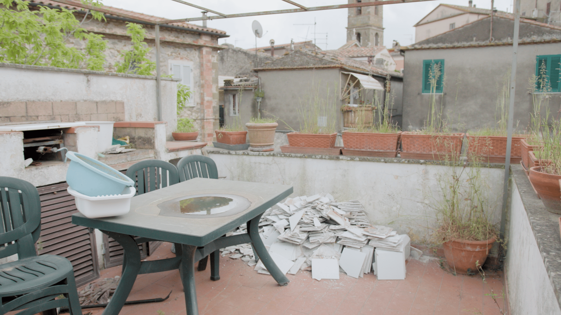 Cluttered rooftop terrace with a plastic table, chairs, cooking area, broken tiles, and overgrown plants.