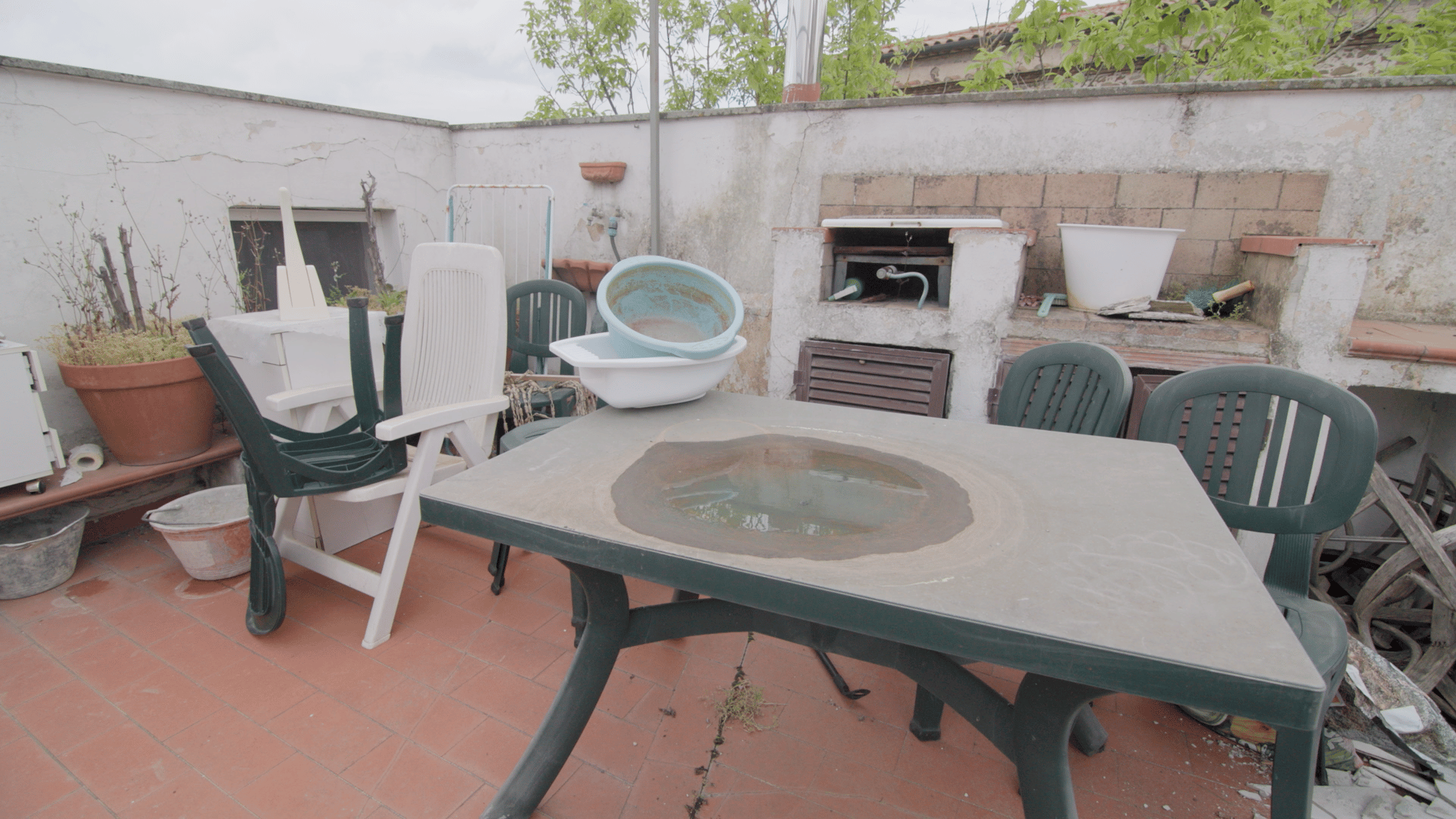 A cluttered rooftop patio with a dirty table, water puddle, plastic chairs, basins, and a built-in barbecue.